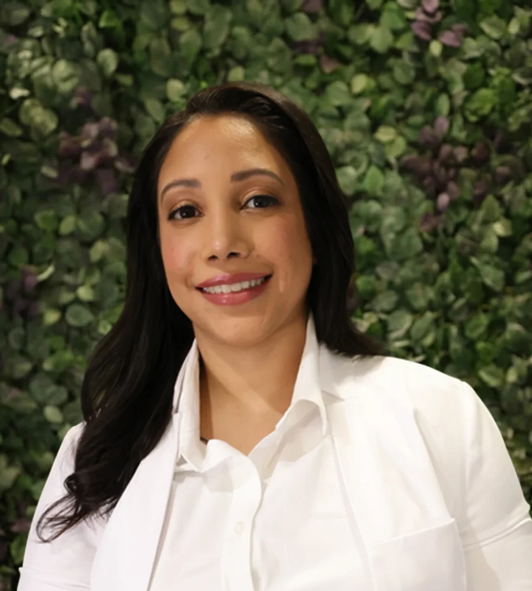 Smiling woman in white shirt, leafy background.